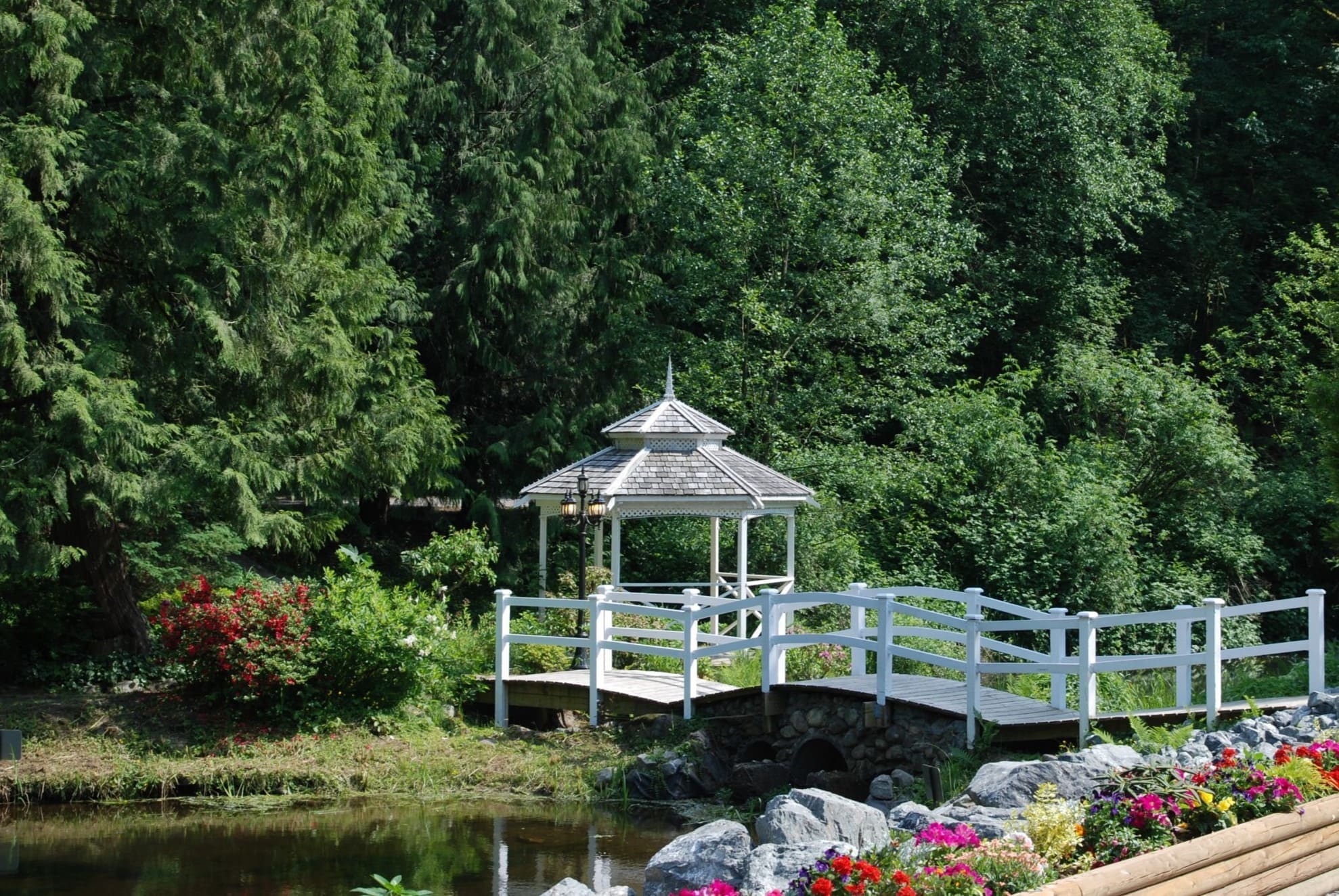 White gazebo and footbridge over a pond surrounded by forest and flowers