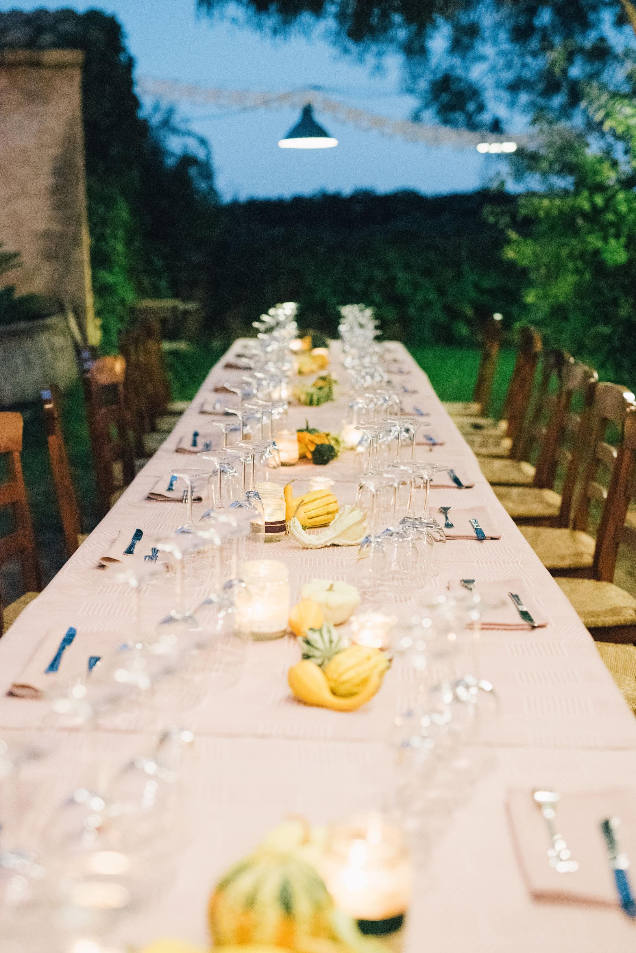A large diverse group seated at a long outdoor dinner table strung with lights beneath the trees