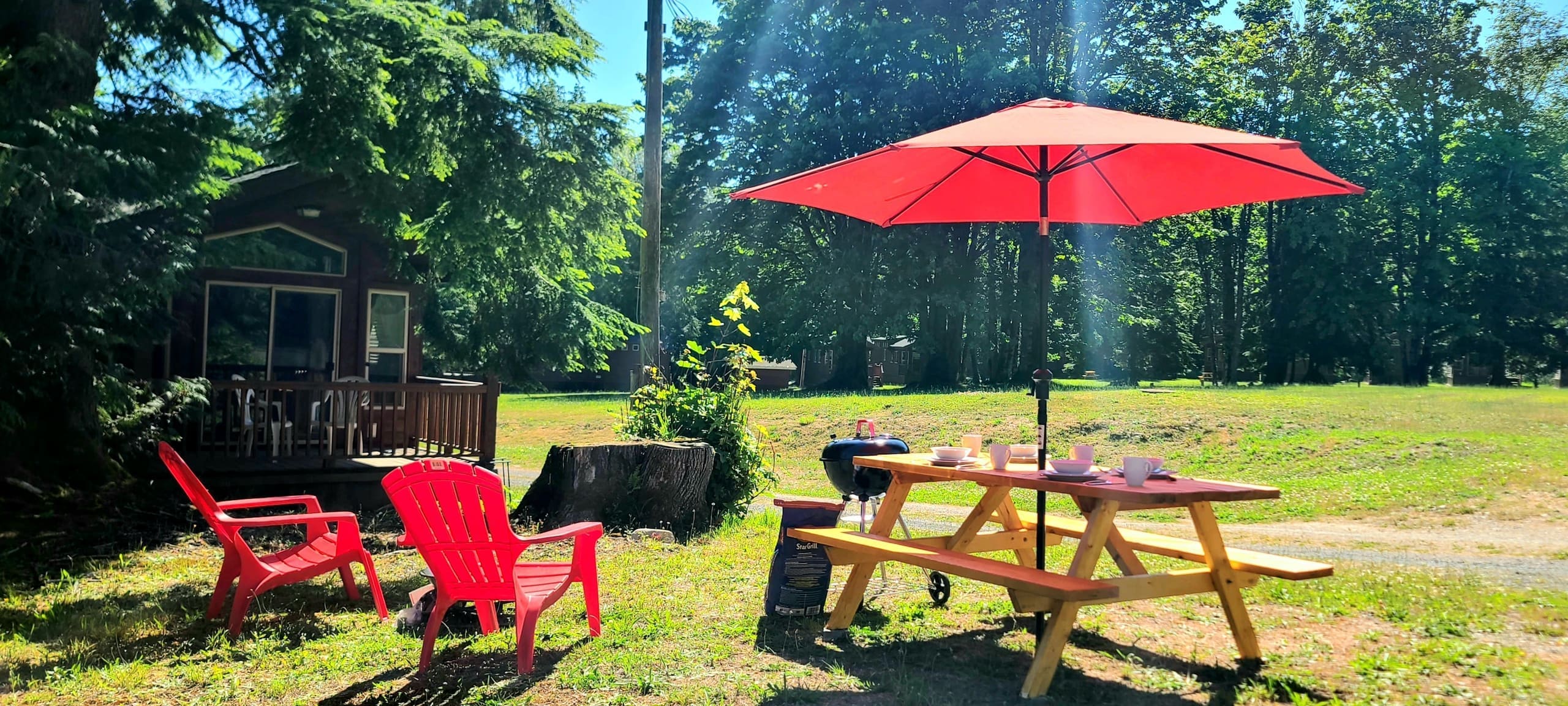 Glamping site with red umbrella, picnic table, and Adirondack chairs by a cabin