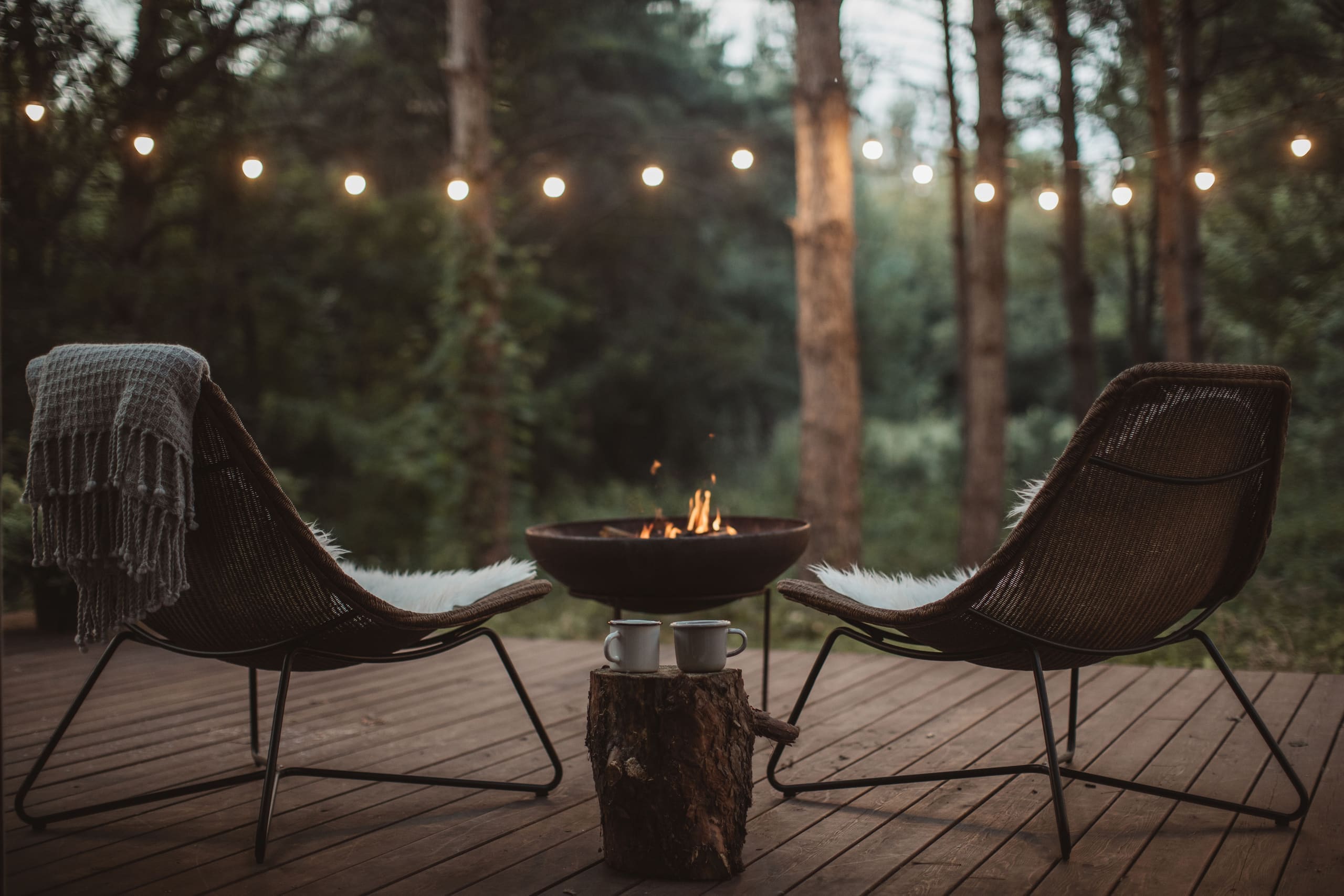 Two chairs and a fire pit on a wooden deck under string lights in the forest at dusk